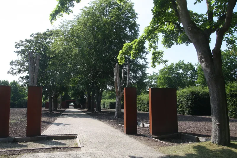 Neben einem gepflasterten Weg stehen rostfarbenen Stelen. Im Kiesbett zu beiden Seiten des Weges werden die Grundrisse der ehemaligen Barackengebäude kenntlich gemacht.