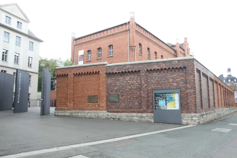 Hinter einer roten Backsteinmauer befindet sich das ehemalige Haftgebäude. An der Mauer ist ein Schaukasten.
