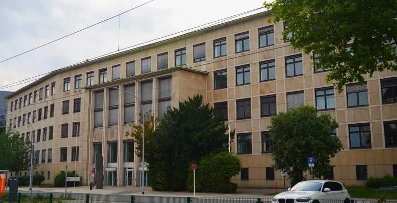 Ein langgestrecktes Gebäude mit vier Stockwerken über einem Kellergeschoss und einem hervortretenden Eingangsbereich ist mit hellen Steinplatten verkleidet.