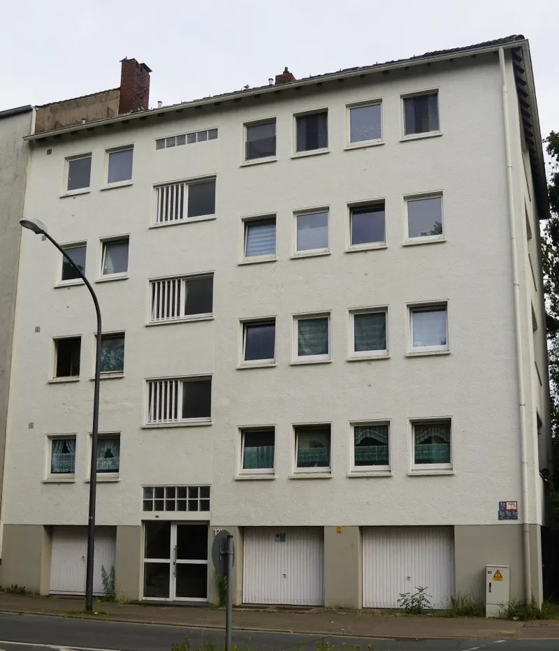 Ein vierstöckiges weißes Wohnhaus an einer Straßenkreuzung. Im Erdgeschoß befinden sich Garagen.