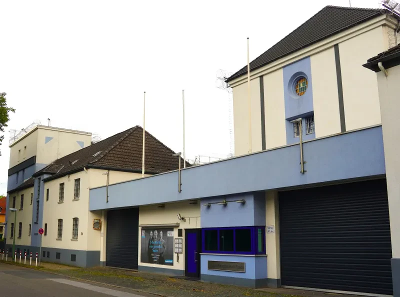 Mehere beige-blaue verputzte Gebäude flankieren den Eingangsbereich mit zwei Rolltoren und einer Sicherheitstür dazwischen.