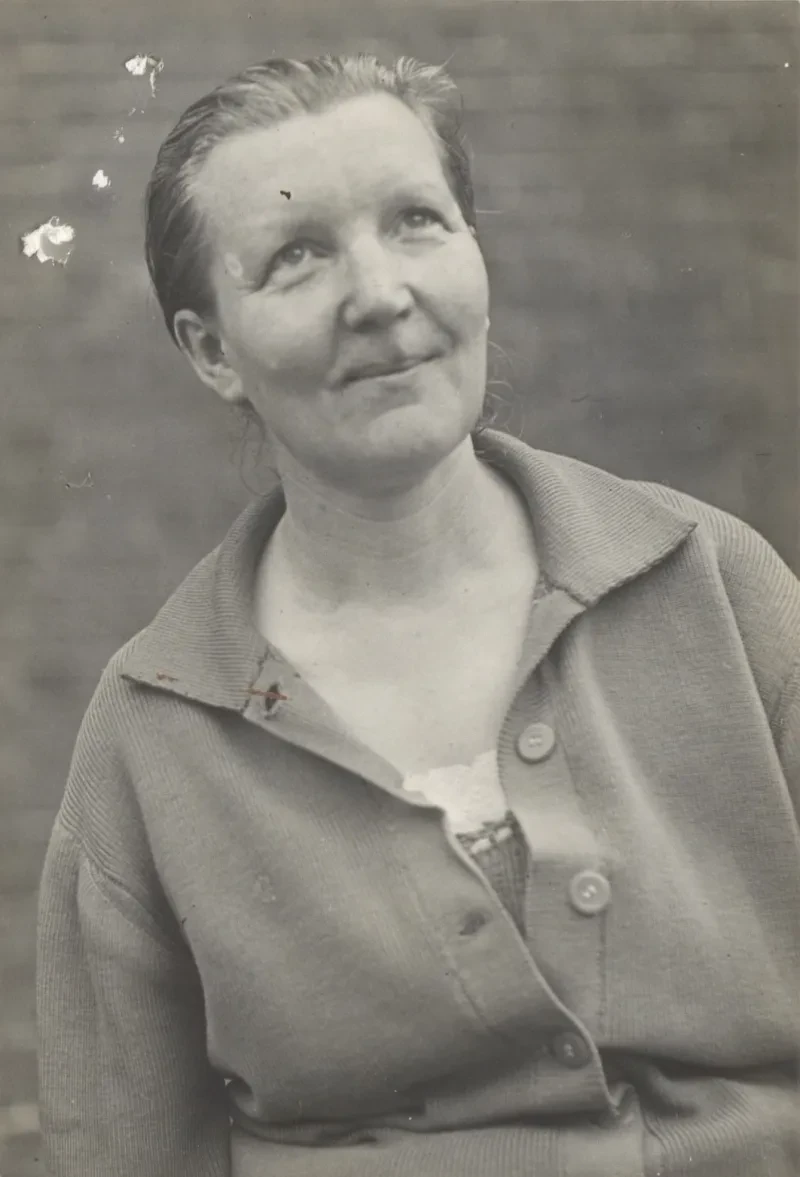 Eine Frau in einer Strickjacke vor einer Mauer. Sie blickt freundlich nach oben.