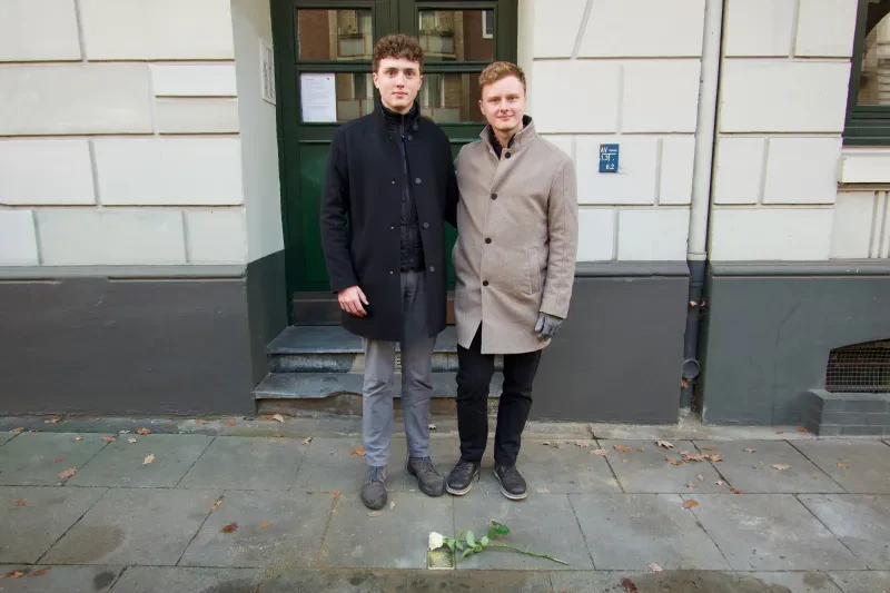 Zwei junge Männer stehen vor einem Stolperstein, dahinter ein Hauseingang.
