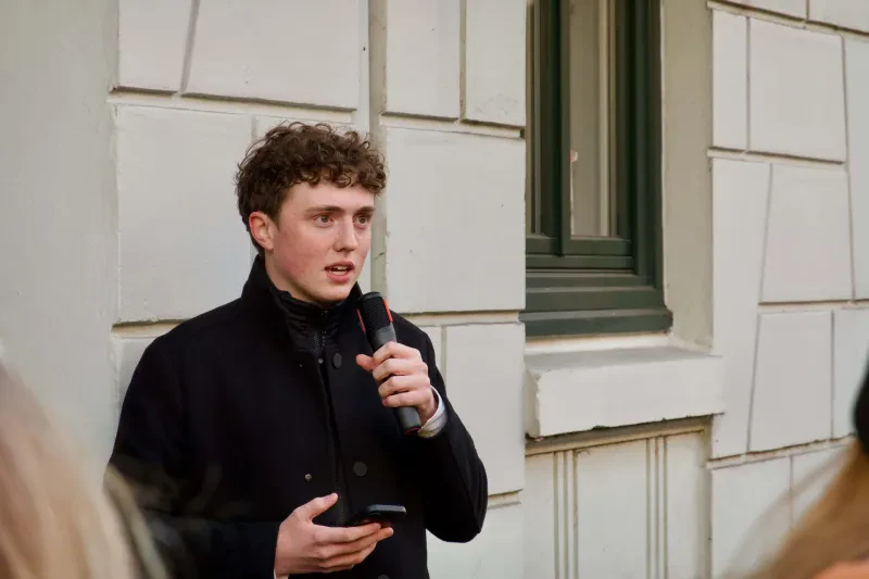 Ein junger Mann steht vor einem Haus und spricht in ein Mikrofon.