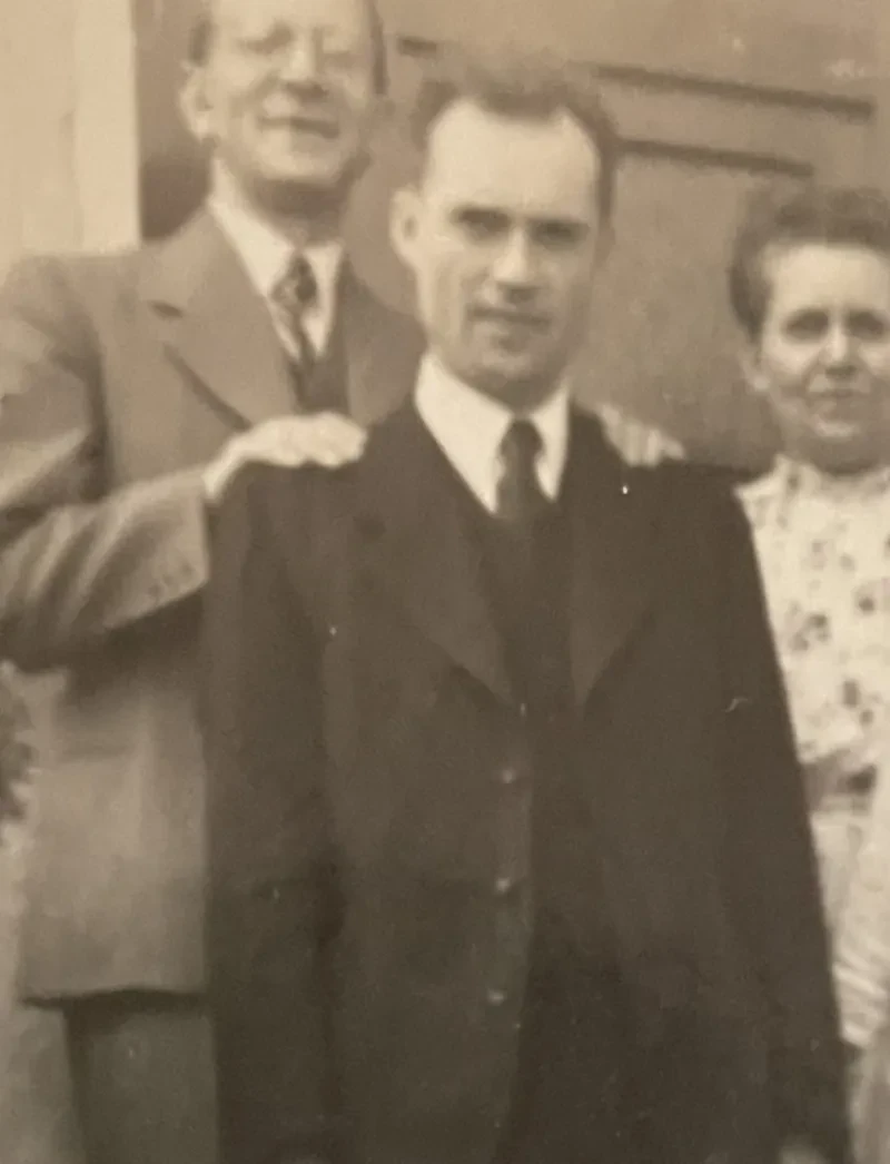Ein junger Mann im Anzug steht vor einem älteren Ehepaar. Der ältere Mann legt seine Hände auf die Schulter des jüngeren Mannes.