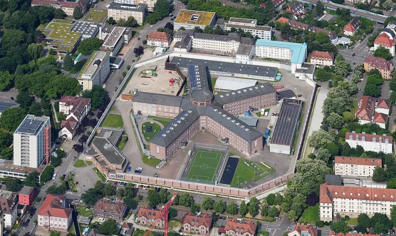 Ein fünfflügeliges Gefängnis ist von einer achteckigen Mauer umgeben. Rundherum stehen Wohnhäuser.