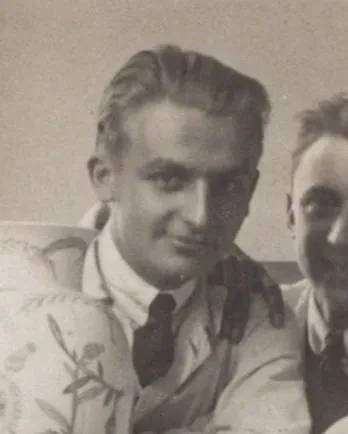 Ein lächelnder junger Mann mit lockigem Haar trägt eine Krawatte und einen Kittel.