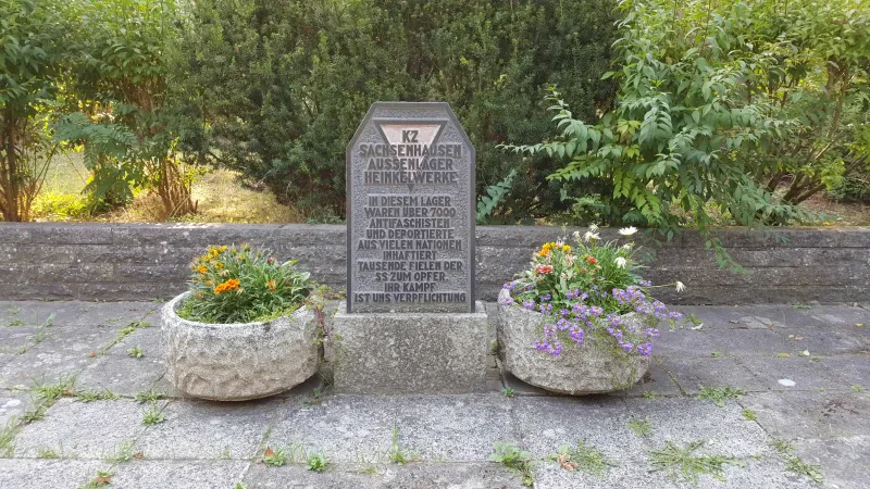 Von zwei Blumenkübeln umrahmter Gedenkstein mit einem roten Winkel und der Aufschrift "KZ Sachsenhausen Aussenlager Heinkelwerke[.] In diesem Lager waren über 7000 Antifaschisten und Deportierte aus vielen Nationen inhaftiert[,] tausende fielen der SS zum Opfer. Ihr Kampf ist uns Verpflichtung[.]"