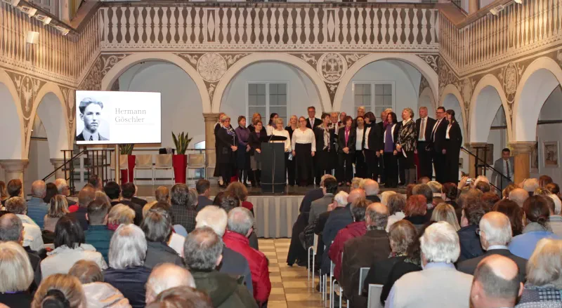 Der mit Menschen gefüllte Rathaushofsaal in St. Veit. Auf der Bühne stehen 25 Männer und Frauen, links von der Bühne ist ein Portrait von Herman Göschler zu sehen.