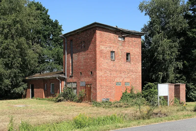 An ein hohes rotes Backsteingebäude mit wenigen, kleinen Fenstern und einer Holztür schließt sich ein kleines einstöckiges Gebäude an.