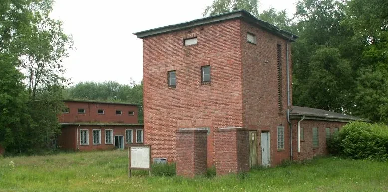 Vor einem roten Trafohaus befindet sich eine Informationstafel. Links von dem Gebäude befindet sich eine weiteres zweistöckiges rotes Backsteingebäude mit einer Fensterreihe.
