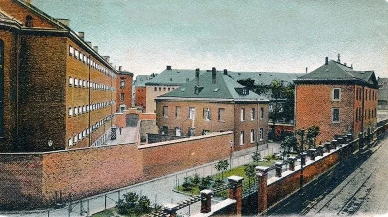 Eine doppelte rote Ziegelmauer trennt ein größeres Areal von einer belebten Straße mit Straßenbahnschienen. Innerhalb der Mauern steht ein langes fünfstöckiges Gefängnisgebäude. Rechts davon stehen zwei weitere zwei- und dreistöckige Gebäude. Alle Gebäude bestehen aus roten Ziegelsteinen.