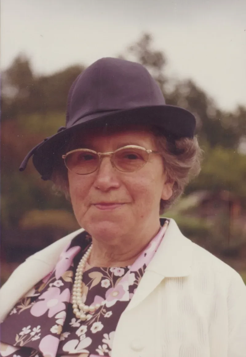 Eine ältere Dame mit lilafarbenem Hut und geblümter Bluse schaut lächelnd zufrieden in die Kamera.