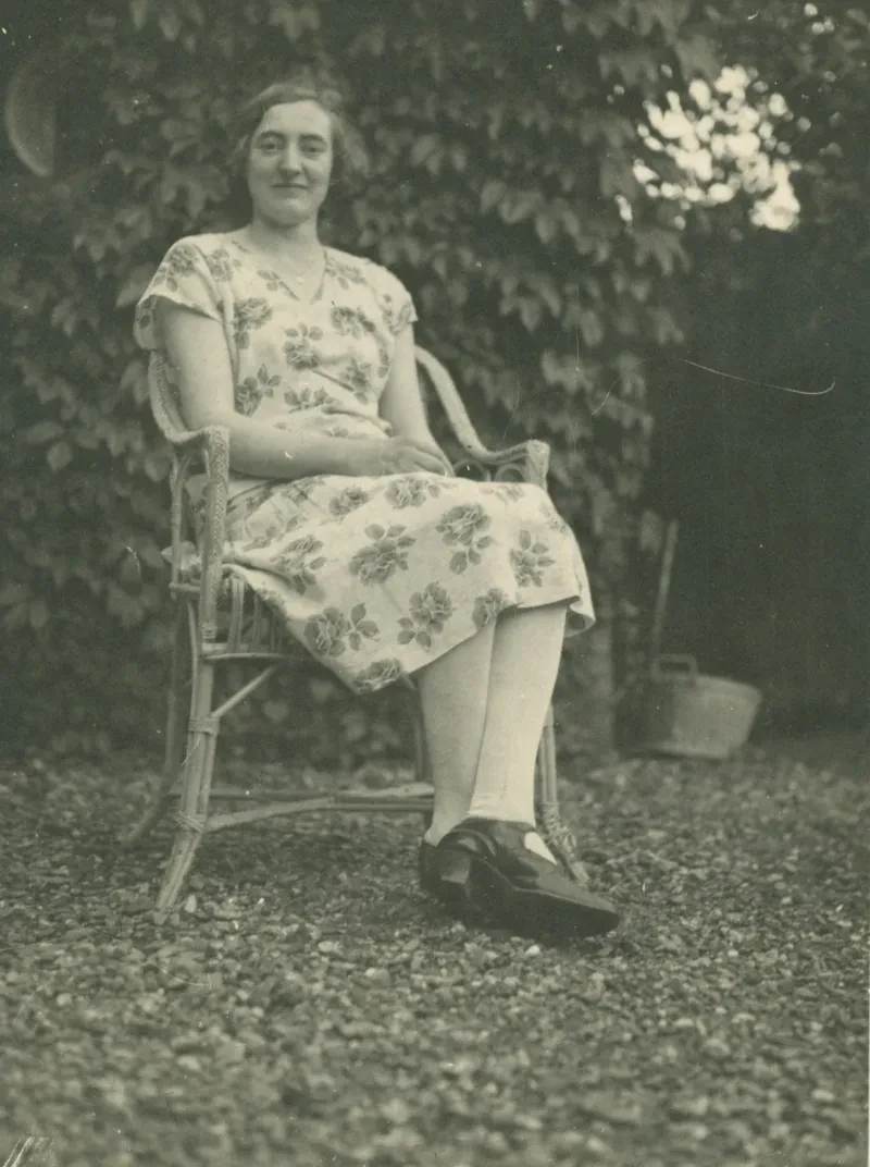 Eine Frau in einem geblümten Kleid sitz in einem Garten oder Park in einem Korbstuhl.