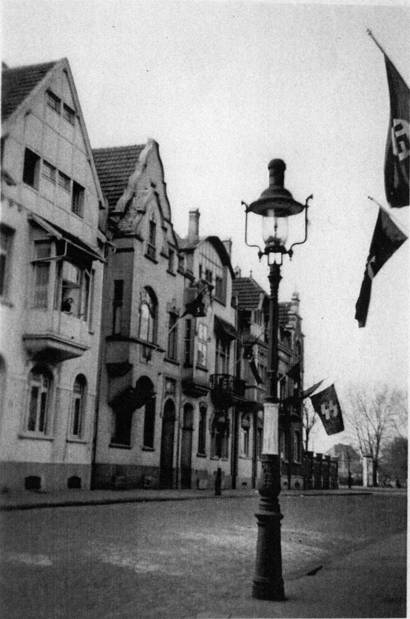 Eine Reihe Häuser, davor ein Laternenpfahl. An den Häusern sind Flaggen mit Hakenkreuzen und der Aufschrift SS gehisst.