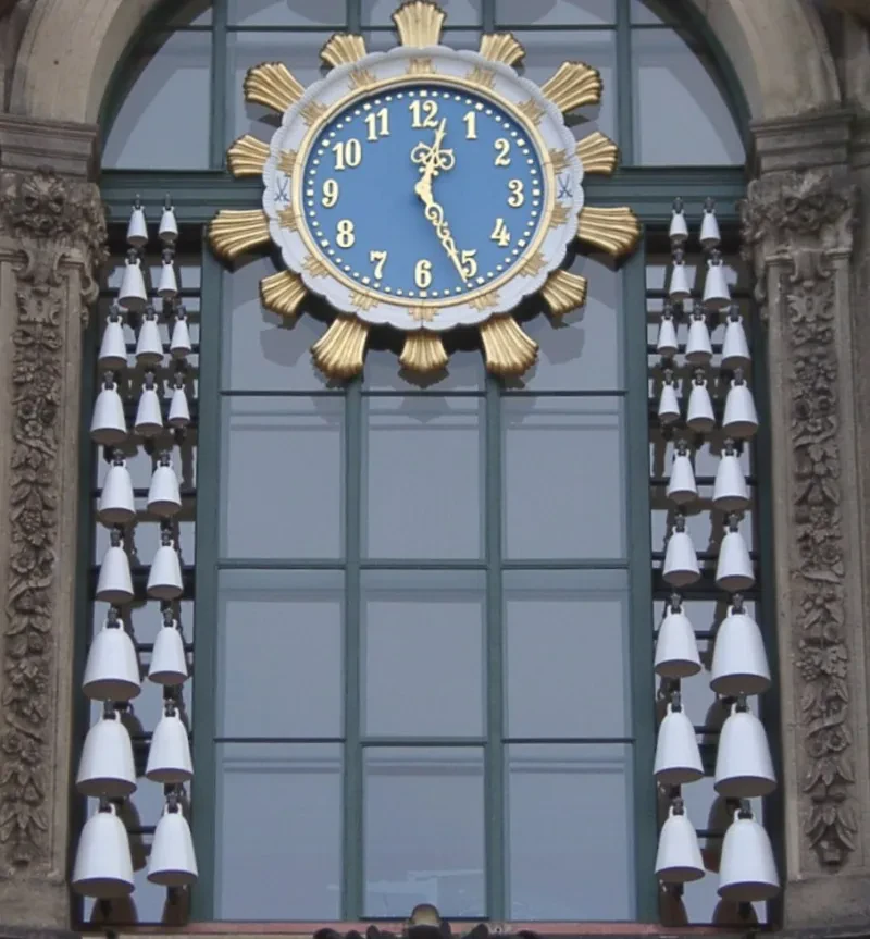 Links und rechts von einer Uhr hängen in einem Fenster je zwanzig weiße Porzellanglocken in unterschiedlicher Größe.