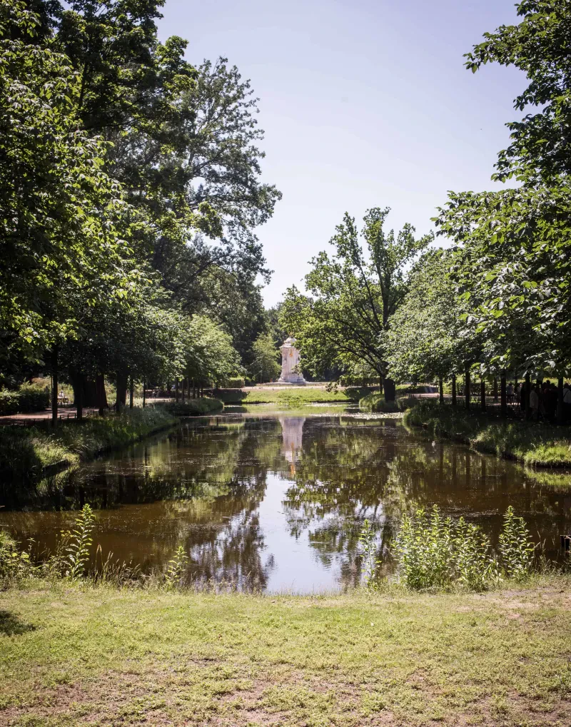 Ein Teich, rechts und links Bäume und im Hintergrund ein Denkmal