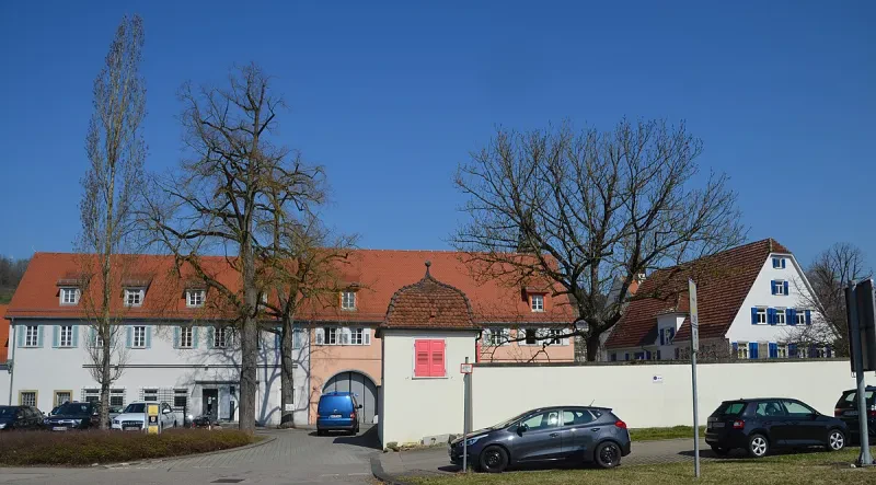Ein beigefarbenes Torgebäude ist von zwei weißen Gebäuden mit mit rotem, ausgebautem Dach umgeben. Vor der Umfassungsmauer befindet sich ein Parkplatz.