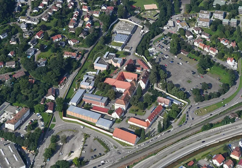 Unregelmäßig angeordnete, meist zwei- oder dreistöckige Gebäude verschiedenen Baustils sind von einer vieleckigen Mauer umgeben. Vor dem Gebäude verlaufen eine Autobahn, eine Landstraße und dazwischen eine Bahnstrecke.
