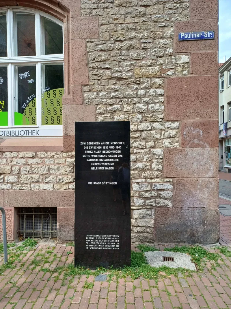 Eine schwarze Gedenktafel mit weißer Schrift vor einem Haus.