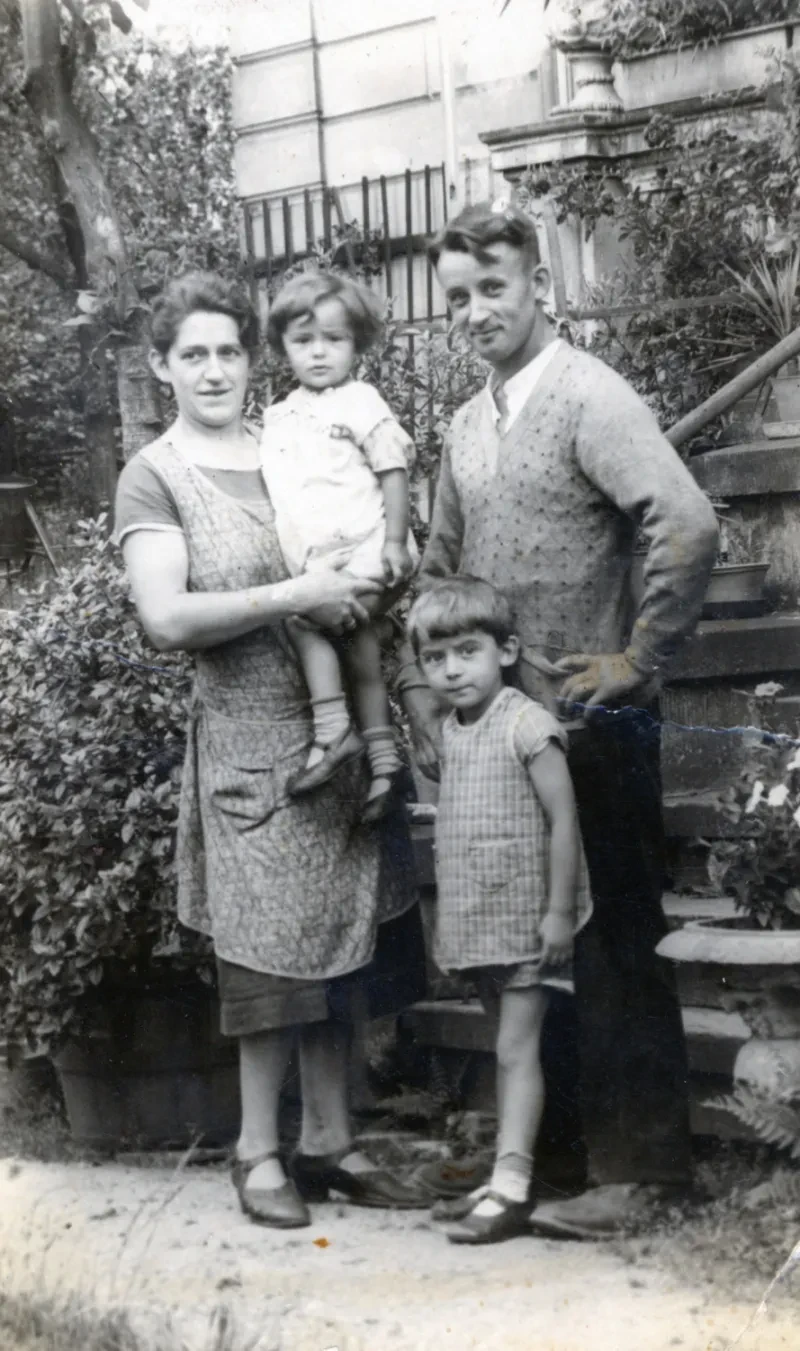 Eine Familie in einem Garten. Die Mutter in einer Schürze trägt die Tochter auf dem Arm. Der Vater fasst den kleinen Jungen an der Schulter.