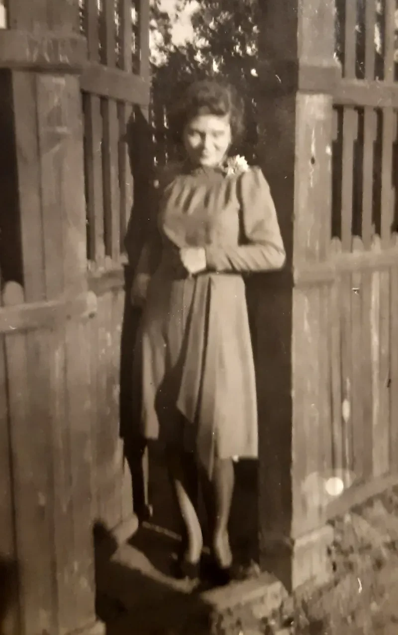 Eine Frau in einem einfarbigen Kleid mit einer Blume auf der Schulter steht vor einem geöffneten Gartentor.
