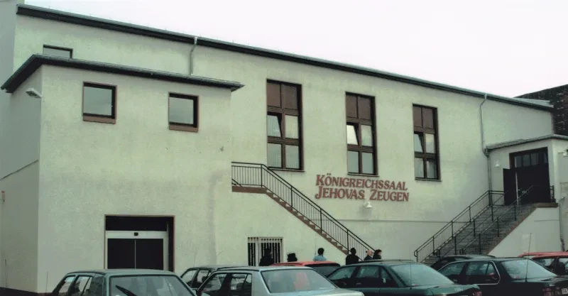 Zwei Freitreppen mit Geländer führen links und rechts zu je einem Vorbau im Obergeschoss eines weiß verputzten Gebäudes in dessen Mitte ein Schild mit der Aufschrift "Königreichssaal Jehovas Zeugen" hängt. Davor befindet sich ein Parkplatz.