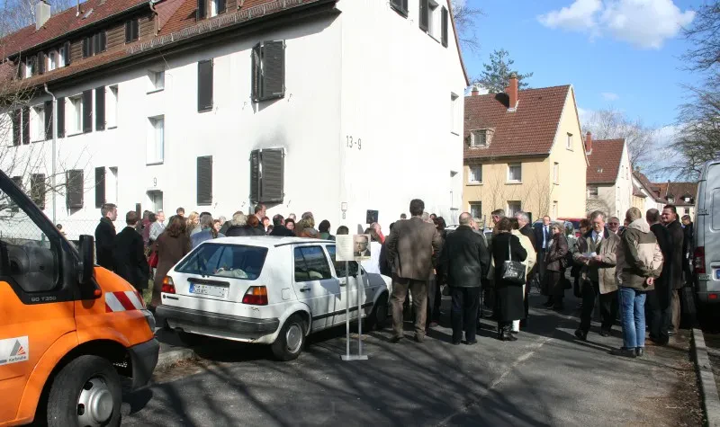 Eine Menschenmenge und einige geparkte Autos vor einem Wohnhaus.