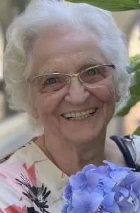 Eine gepflegt aussehende grauhaarige Frau im Sommerkleid mit Brille und Blumen in der Hand lächelt in die Kamera.