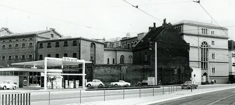 Hinter einer Tankstelle sieht man auf einem Schwarz-Weiß-Foto eine hohe Backsteinmauer. Darüber hinaus ragen einige drei- und vierstöckige Gebäude, die miteinander verbunden sind.