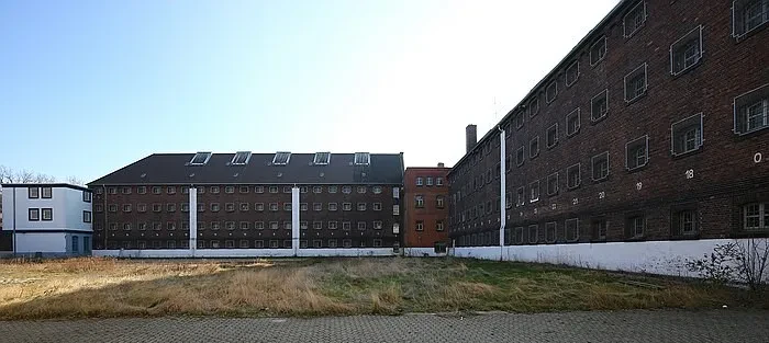 Zwei rotbraune vierstöckige Zellengebäude sind im rechten Winkel miteinander verbunden. Im Hof befindet sich eine ungeplegte Wiese.