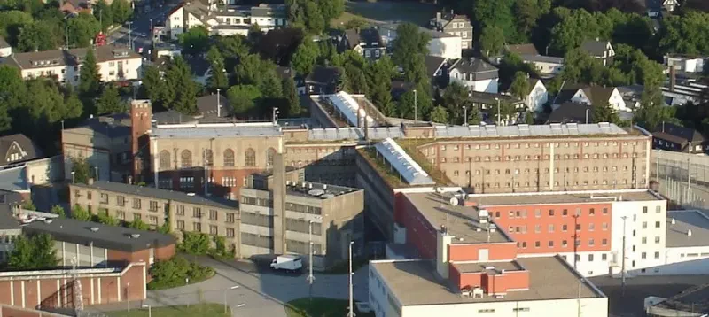 Auf der Luftaufnahme sieht man inmitten eines Dorfes die ummauerte Anlage eines Gefängnisses. Neben einem Neubau steht das ursprüngliche vierflügelige, vierstöckige Gebäude.