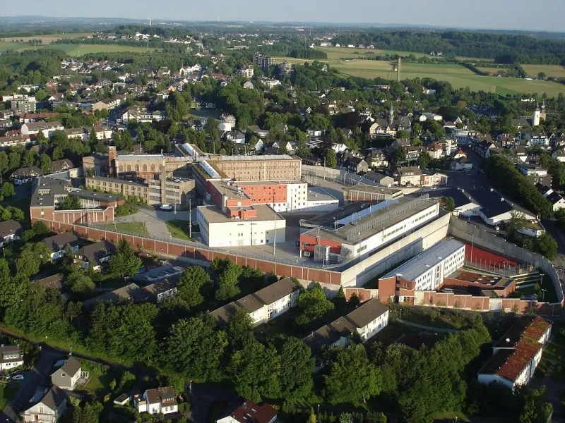 Auf der Luftaufnahme sieht man inmitten eines Dorfes die ummauerte Anlage eines Gefängnisses. Neben einem Neubau steht das ursprüngliche vierflügelige, vierstöckige Gebäude.