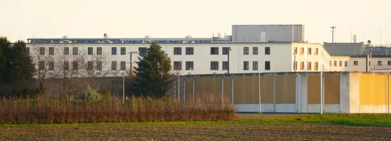 HInter einem Acker verdeckt eine hohe, gelbe Mauer teilweise die Sicht auf ein langgestrecktes weißes, vierstöckiges Gebäude.