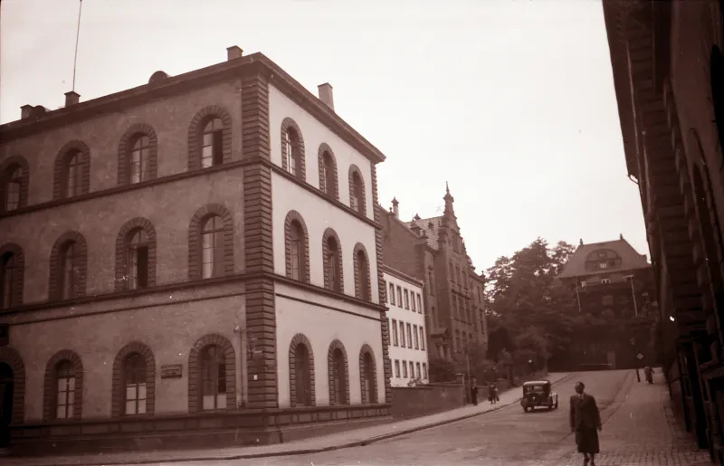 Vorne links das Landgerichtsgebäude Kaiserslautern, rechts davon eine Straße mit Fußgängern und einem fahrenden Auto.