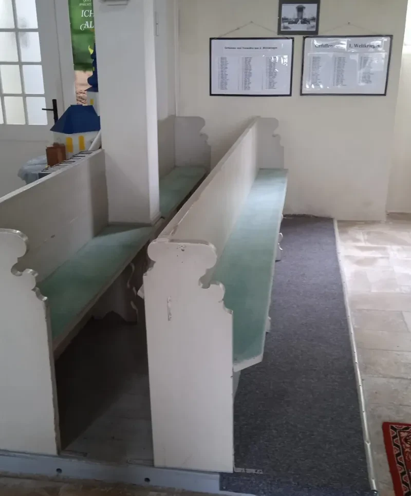 Neben Kirchenbänken befinden sich an einer Wand zwei gerahmte Tafeln mit Namenslisten in je drei Spalten.
