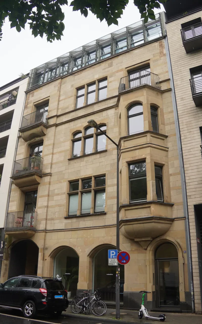 Auf dem Flachdach des ursprünglich vierstöckigen, hellgelben Wohnhaus wurde später noch ein Stockwerk mit Glasfassade aufgesetzt. Im Erdgeschoss befindet sich ein Laden.