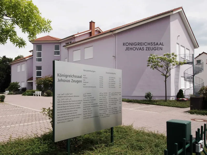Ein Gebäude mit einem Schild davor. Auf dem Gebäude und dem Schild steht "Königreichssaal Jehovas Zeugen". Auf dem Schild sind mehrsprachig die Versammlungszeiten aufgeführt.