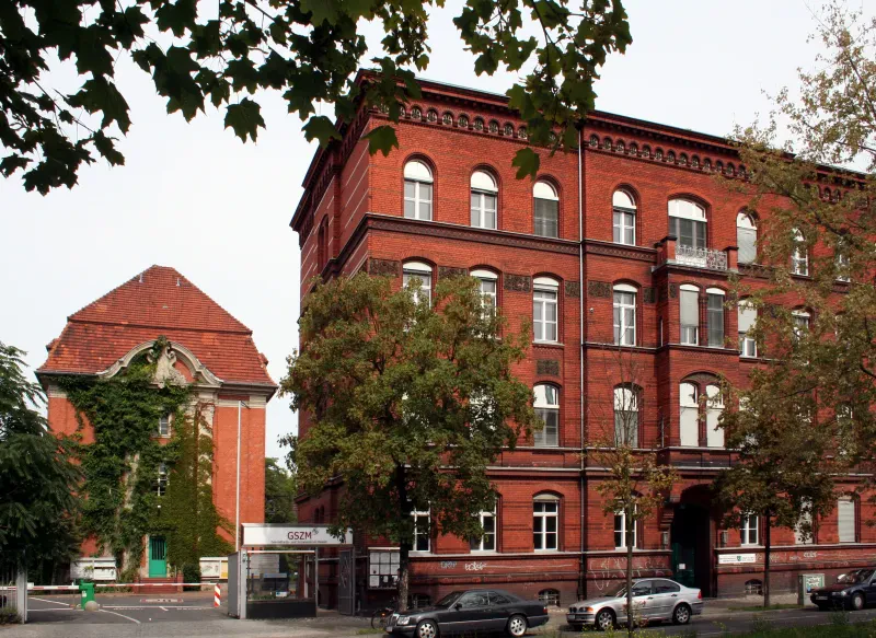 Zwei mehrstöckige rote Ziegelgebäude, davor parken drei Autos. Links der Eingang des Krankenhausgeländes mit einer Schranke.