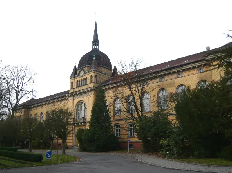 Ein zweigeschossiges langes Gebäude mit Bäumen und Büschen davor. Über dem Portal befindet sich eine Kuppel mit einer Uhr.