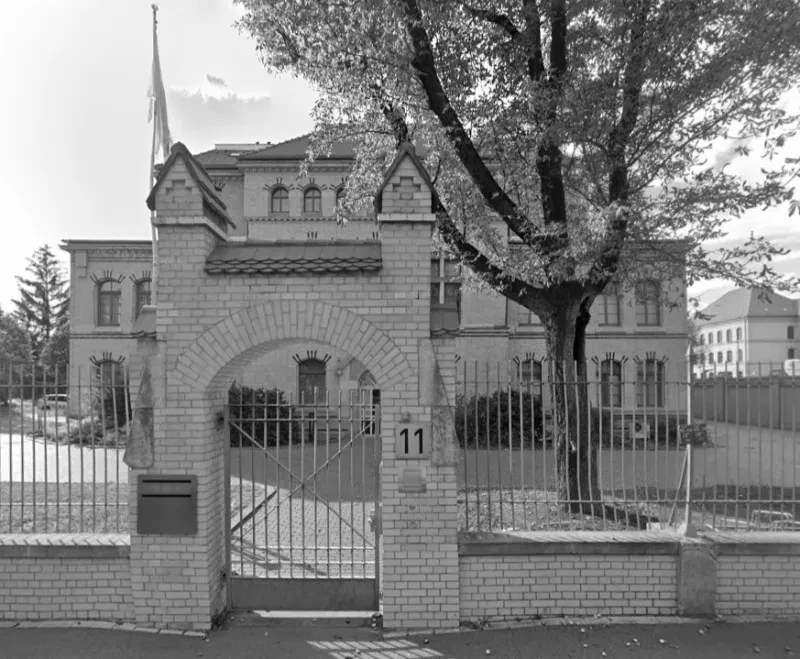 Schwarzweißfoto eines Gitterzauns, dahinter ein villenartiges Gebäude.
