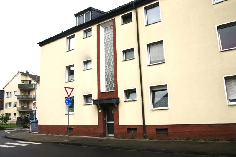 Ein dreistöckiges, hell verputztes Wohnhaus steht an einer Straßenkreuzung.