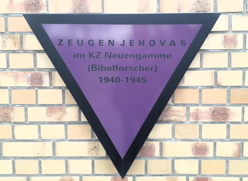 Ein lila Winkel an einer Mauer trägt die Schrift: Zeugen Jehovas im KZ Neuengamme (Bibelforscher) 1940 - 1945