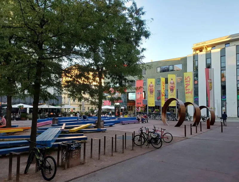 Ein Platz mit Bäumen und bunten Bänken, vorne stehen Fahrräder, rechts hinten sind drei große Ringe zu sehen, die das Denkmal bilden.