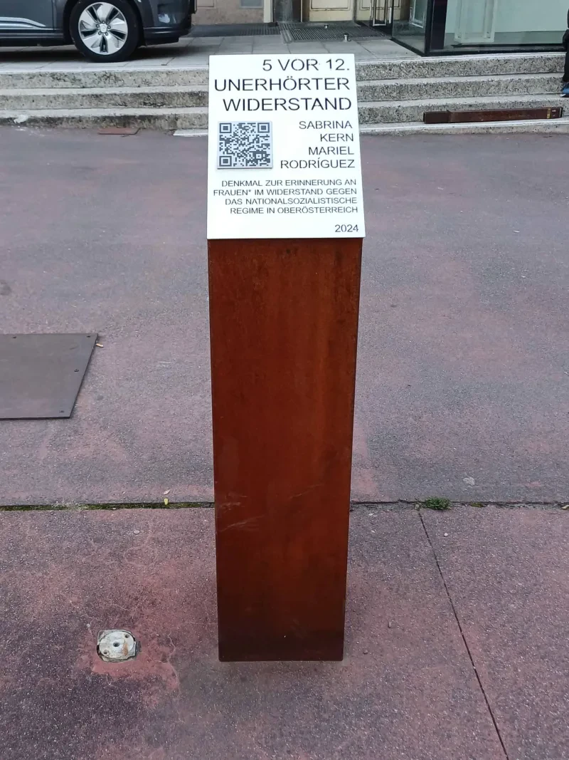 Stele mit einem Schild oben und der Inschrift „5 vor 12. Unerhörter Widerstand. Sabrina Kern [,] Mariel Rodríges [.] Denkmal zur Erinnerung an Frauen* im Widerstand gegen das nationalsozialistische Regime in Oberösterreich [,] 2024“