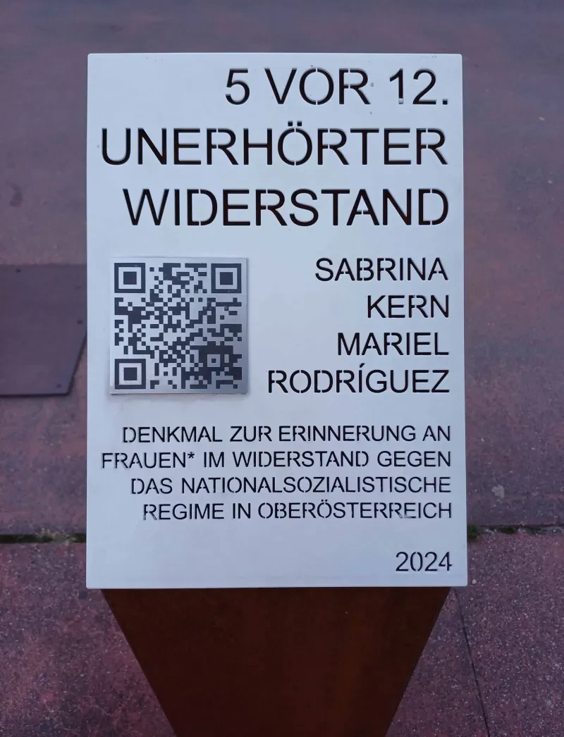Schild auf der Stele mit der Inschrift „5 vor 12. Unerhörter Widerstand. Sabrina Kern [,] Mariel Rodríges [.] Denkmal zur Erinnerung an Frauen* im Widerstand gegen das nationalsozialistische Regime in Oberösterreich [,] 2024“