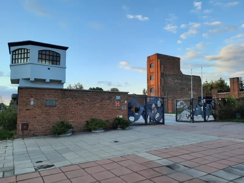 Auf einer roten Backsteinmauer befindet sich ein weißer Wachturm. Hinter dem Eingang steht die vierstöckige Ruine eines ausgebrannten ehemaligen Gefängnisses in einem Fabrikgebäude.