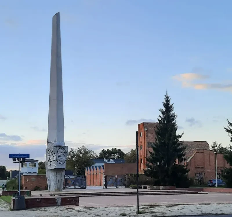 An einer Straßenkreuzung steht ein hoher, weißer Obelisk. Dahinter sieht man die aus roten Backsteinen erbaute Ruine eines ehemaligen Gefängnisses.