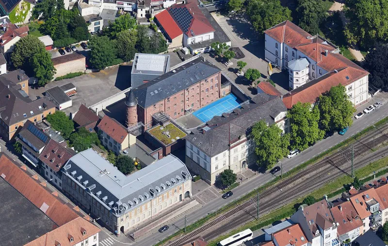 HInter einer Bahnlinie und einer Stadtstraße befindet sich ein helles dreistöckiges Gerichtsgebäude. Dahinter befindet sich von einer Mauer umgeben ein rotes, vierstöckiges Backsteingebäude, ein Turm und ein Tennisplatz.