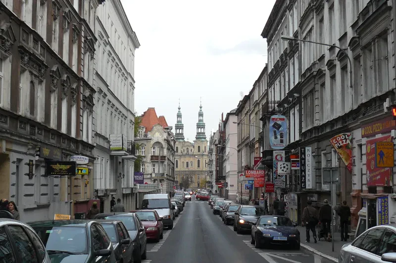 Ein Blick in eine Straße einer Großstadt mit Fußgängern und parkenden Autos. Im Hintergrund ist eine zweitürmige Kirche zu sehen.